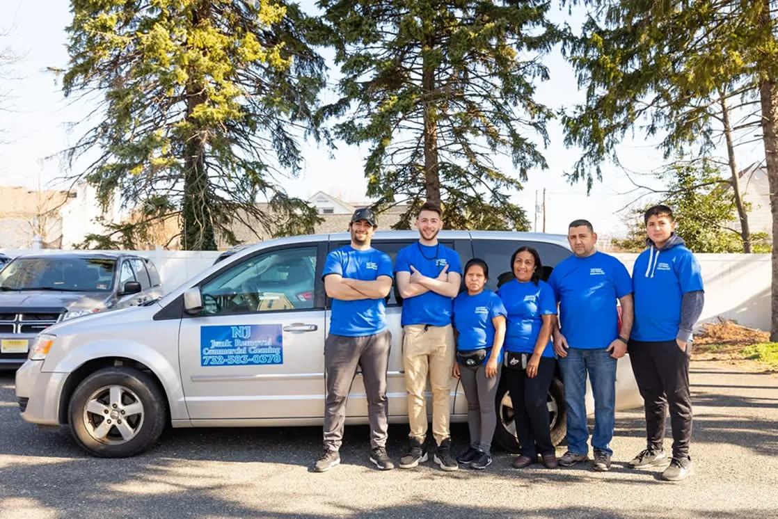 The six-person NJ Indoor Outdoor crew in blue company T-shirts standing in front of their silver minivan, tall pines behind them