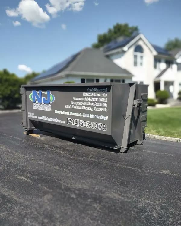 A grey NJ Indoor Outdoor roll-off dumpster sitting on the driveway of a white suburban house under a blue sky