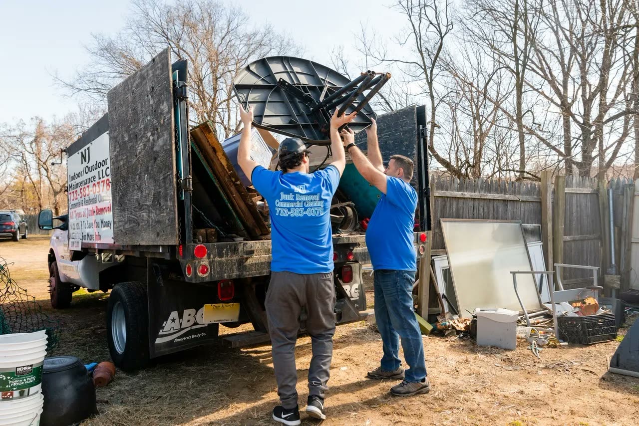 Two men in blue company T-shirts lifting a black patio table over the side of a loaded dump truck in a back yard