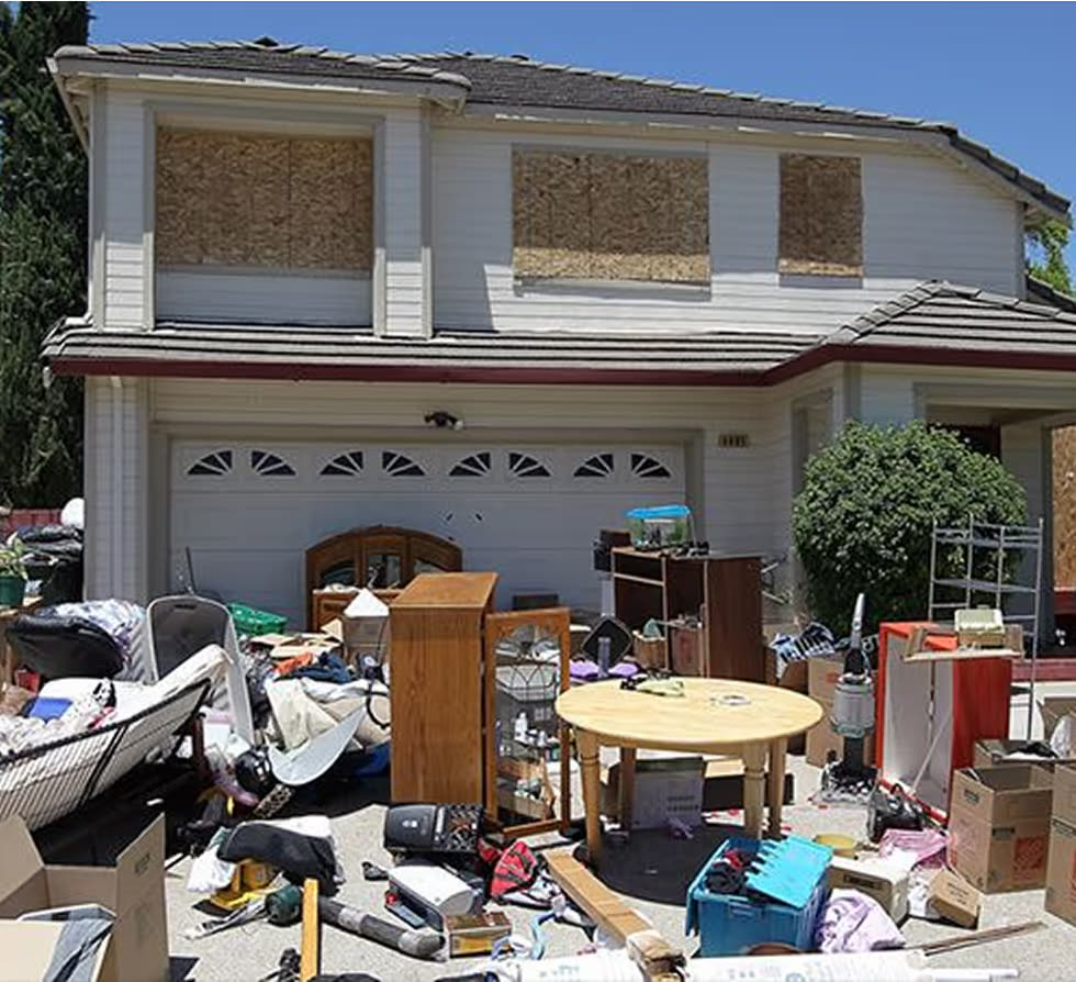 A two-storey house with boarded windows, its driveway and lawn buried under furniture, boxes and luggage