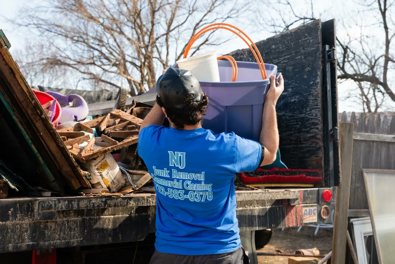 A worker seen from behind hoisting a purple storage tote and buckets up into a loaded dump truck