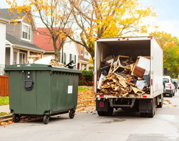 A box truck loaded with old furniture and debris beside a waste bin on a residential street